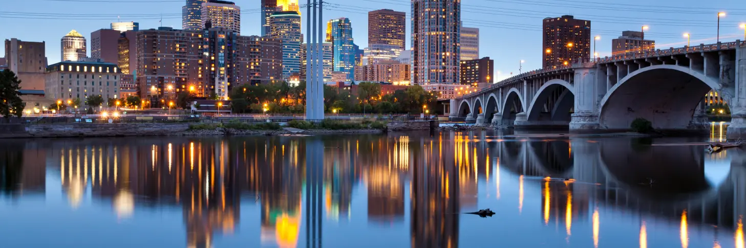 City skyline at dusk with illuminated buildings reflecting on a calm river; a bridge is visible on the right.
